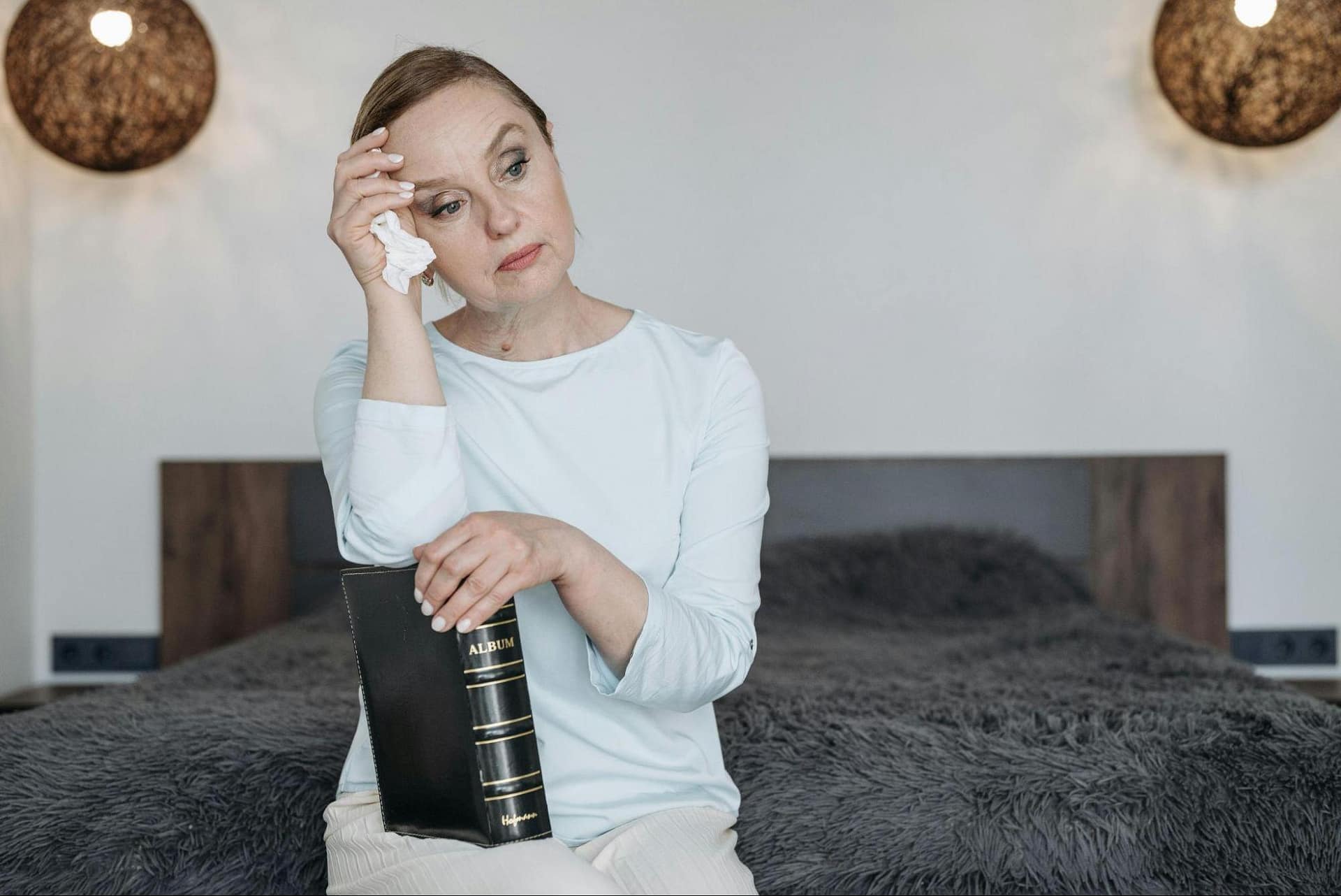 A woman sitting on a bed holding a photo album, looking emotional as she reflects on memories.