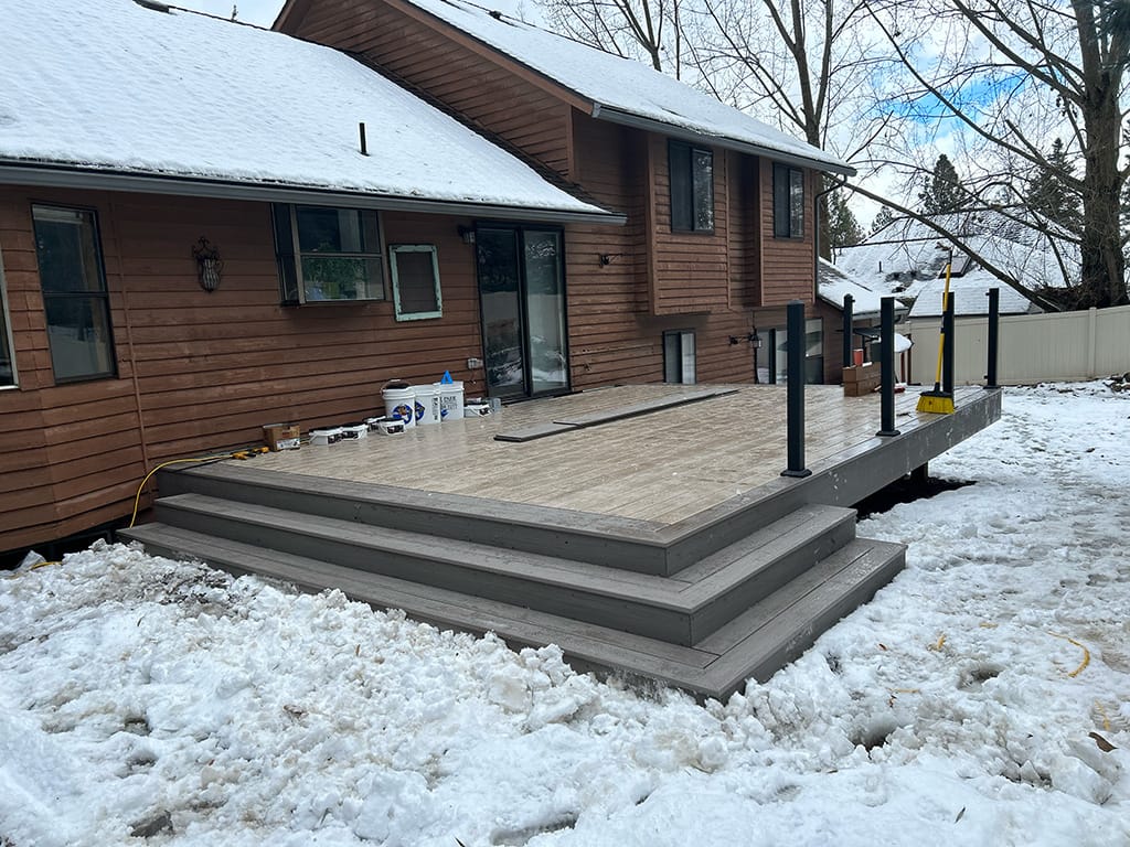 A deck under construction with TimberTech AZEK PVC decking and a Fortress Fe26 3" handrail. Tools are scattered on the wet deck surface, surrounded by snow on the ground and on the roof of the house.