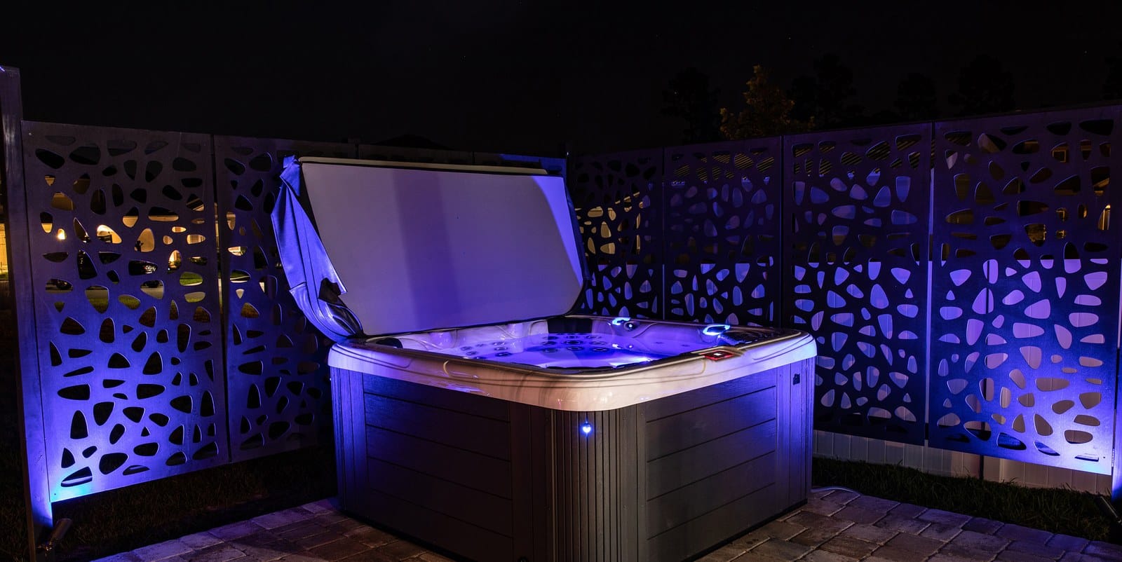 A Garden Leisure hot tub set on a paver patio at night, its cover held open on a cover lifter