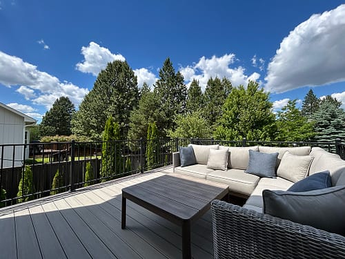 Spacious deck featuring L-shaped furniture and a coffee table, set against lush greenery and a serene sky.