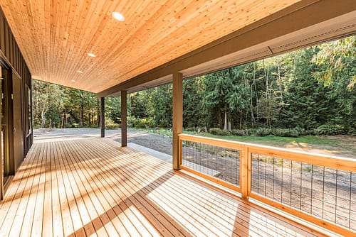 Ground-level wooden deck in the Pacific Northwest with a modern roof, tongue-and-groove soffit, wooden handrails, and metal wire accents.