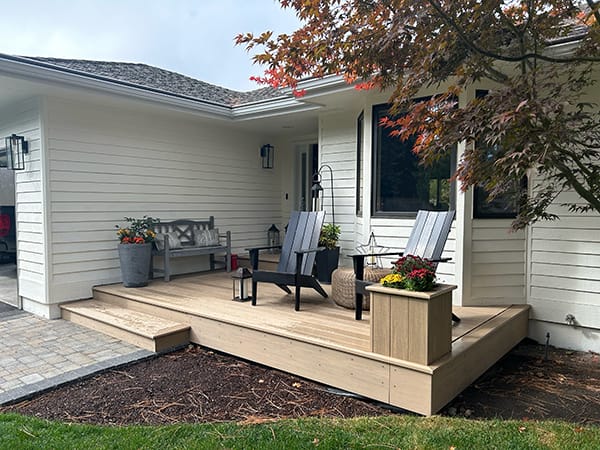 The front porch of the home is constructed with weathered teak decking, featuring a single step up from a paver patio walkway. A planter box positioned at the corner of the porch complements the deck, enhancing its aesthetic appeal.