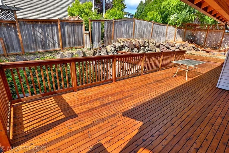 Large cedar deck with an orange stain in a backyard, facing a fence with a small rock wall, green trees, and a neighboring house in the background.