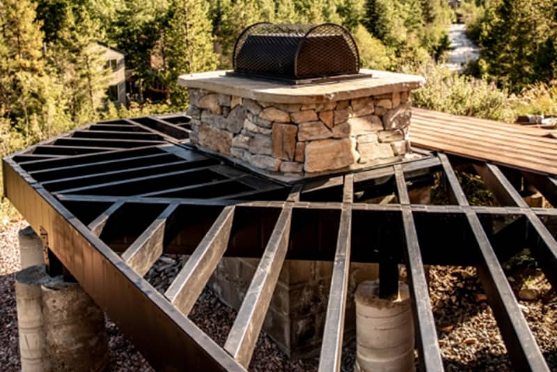 Steel-framed deck using Fortress Evolution framing, with a rock-built grill station and green trees in the background.