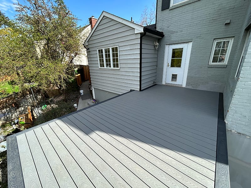 Gray deck with a darker picture frame facing a brick house, featuring a staircase and a green tree on the left side.