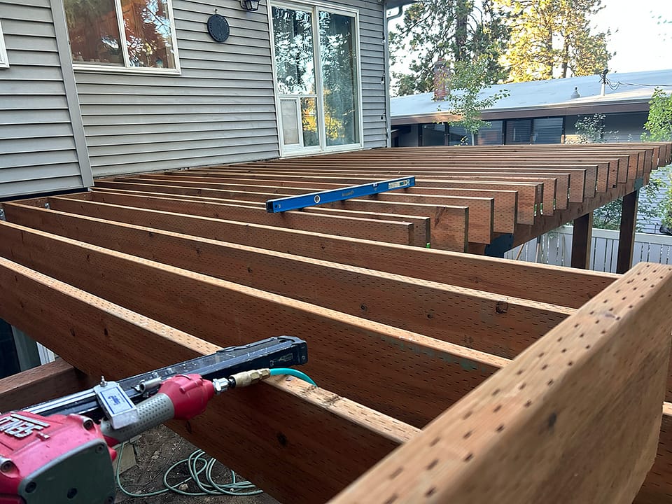 Partially framed deck with a nail gun, blue 4-foot level, and pressure-treated lumber in a backyard, 9 feet off the ground.