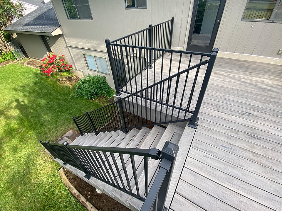 View from the top of a completed deck staircase with handrail, decking, and fascia, leading down to a paver landing in a green backyard with red roses to the right.