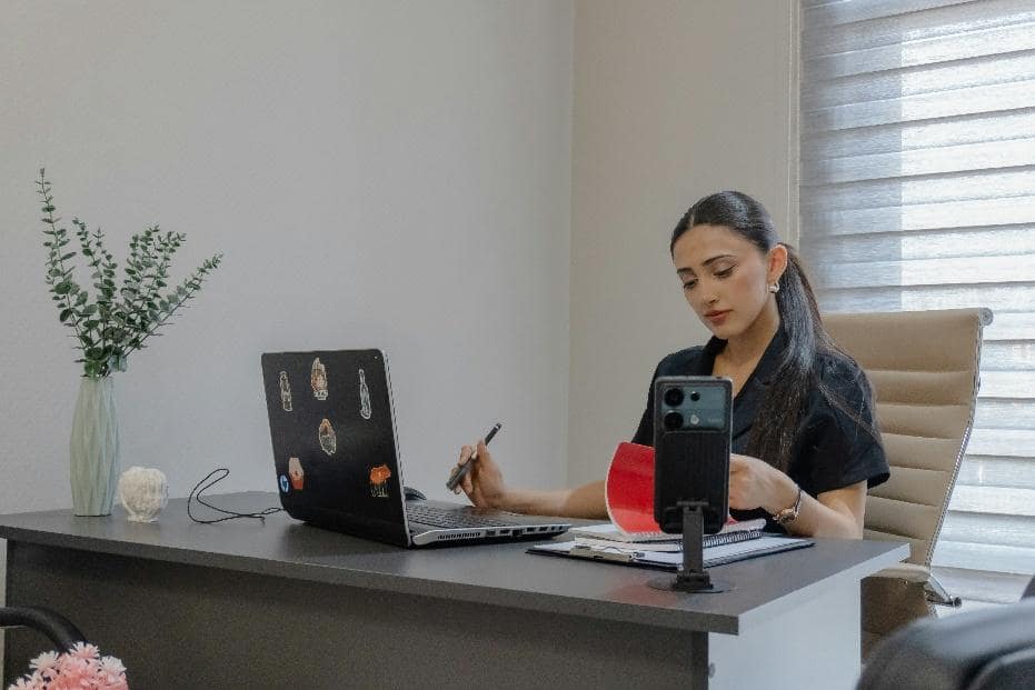 Healthcare professional at a desk using a phone on a stand and laptop.